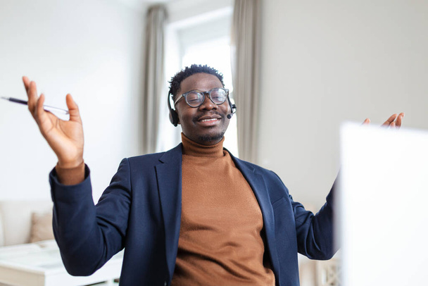 Sourire employé afro-américain dans les écouteurs à l'aide d'un ordinateur portable, regarder l'écran, faire un appel vidéo ou regarder webinaire, notes d'écriture, concept de langue d'apprentissage à distance, opérateur de centre d'appels travaillant - Photo, image