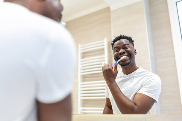 Homem africano sorridente com escova de dentes limpando os dentes e olhando espelho no banheiro. Um jovem bonito a escovar os dentes de manhã na casa de banho. Cara feliz de pijama escovando os dentes à noite antes de ir dormir. - Foto, Imagem