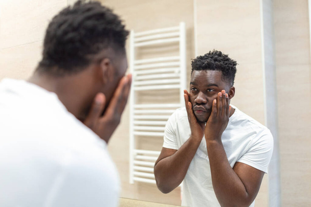 Igiene mattutina, bell'uomo in bagno che si guarda allo specchio. Riflessione di Africanman con la barba guardando specchio e toccando il viso in bagno governare - Foto, immagini
