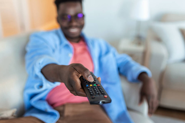 Aufgeregter Afroamerikaner zeigt Fernsehkontrolleur auf Kamera, die Kanäle vor dem Fernseher schaltet. Werbung für Fernsehprogramme. Selektiver Fokus - Foto, Bild