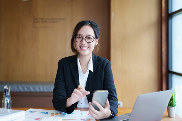 Muotokuva nuori aasialainen nainen näyttää hymyilevät kasvot, kun hän käyttää puhelinta, tietokonetta ja taloudellisia asiakirjoja pöydällään aikaisin aamulla. - Valokuva, kuva