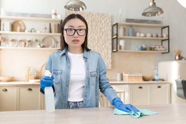 Portrait of a young Asian housewife cleaning at home. Wipes the table in the kitchen with a rag in rubber gloves, uses household chemicals. He is standing, holding a spray, looking at the camera. - Zdjęcie, obraz