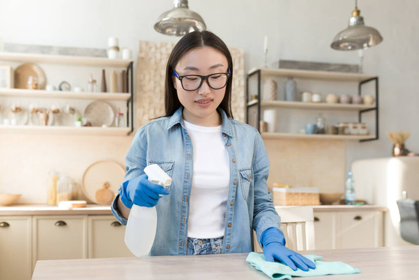 Maid services. Young beautiful Asian woman works as a cleaner. Cleans the house. Wipes the table with a cloth in rubber gloves, sprays the surface with a spray. - Foto, immagini