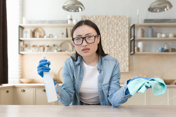Tired young beautiful Asian woman cleaning at home. Wipes the table with a rag in rubber gloves and a bottle of spray. He looks at the camera, spreads his hands. - Φωτογραφία, εικόνα