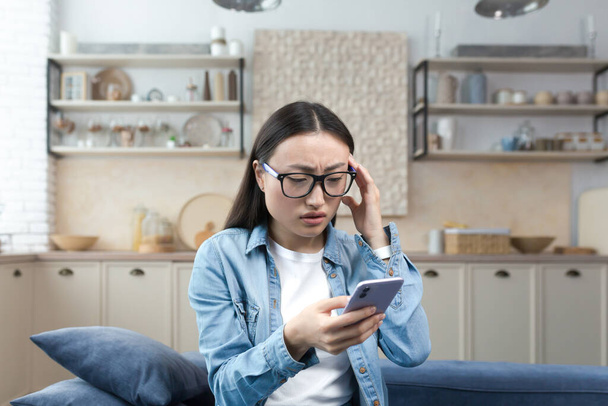 Worried young Asian woman holds the phone in her hands, received bad news by message. Sitting at home on the sofa, holding her head, nervous. - Foto, Imagen