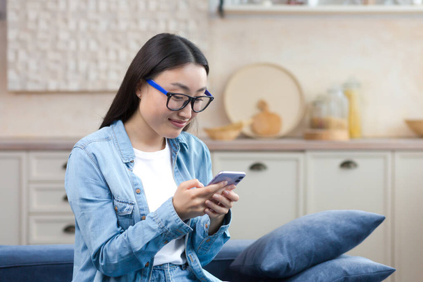 A happy young beautiful Asian woman is sitting on the sofa at home. She is holding a mobile phone in her hands, writing a message. Corresponds with boyfriend, friends, smiles - 写真・画像