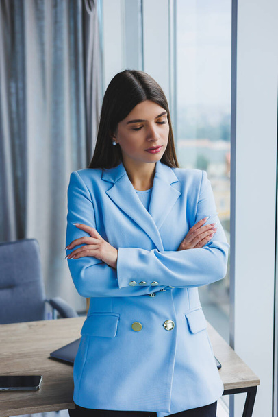 Portrait of a pretty brunette in a blue jacket in the office near the panoramic window with a view of the city. Happy business woman in the office - Photo, image