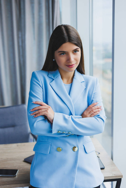 Portrait of a pretty brunette in a blue jacket in the office near the panoramic window with a view of the city. Happy business woman in the office - Фото, изображение
