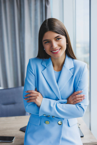 Ofisteki panoramik pencerenin yanında şehir manzaralı mavi ceketli güzel bir esmerin portresi. Ofisteki mutlu iş kadını. - Fotoğraf, Görsel