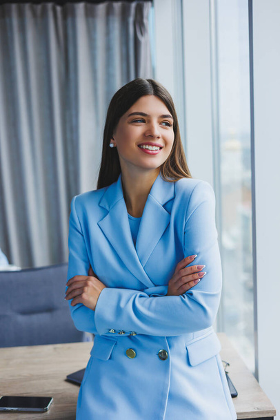 Portrait of a pretty brunette in a blue jacket in the office near the panoramic window with a view of the city. Happy business woman in the office - Foto, imagen