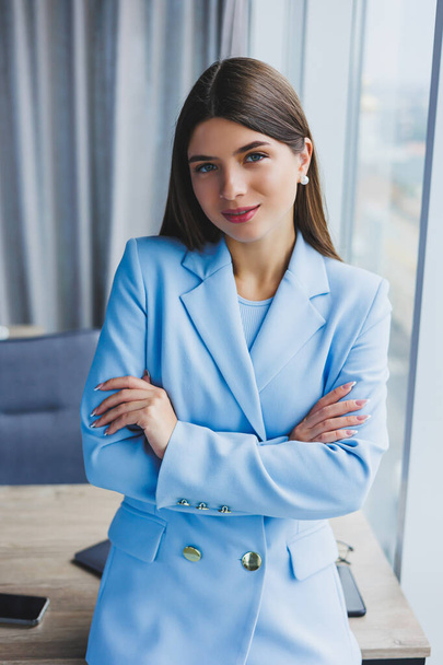 Portrait of a pretty brunette in a blue jacket in the office near the panoramic window with a view of the city. Happy business woman in the office - Fotó, kép