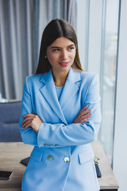 Portrait of a pretty brunette in a blue jacket in the office near the panoramic window with a view of the city. Happy business woman in the office - Valokuva, kuva