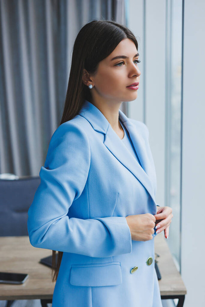 Portrét krásné brunetky v modré bundě v kanceláři poblíž panoramatického okna s výhledem na město. Šťastná obchodnice v kanceláři - Fotografie, Obrázek