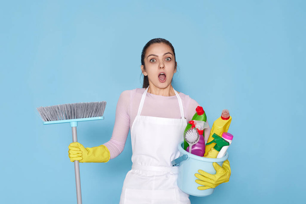 surprised young woman in rubber gloves and cleaner apron holding bucket of detergents and broom on blue background. - Foto, Imagen