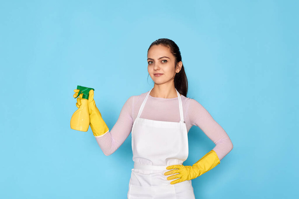 young confident woman in rubber gloves and cleaner apron holding detergent sprayer on blue background. - Fotografie, Obrázek