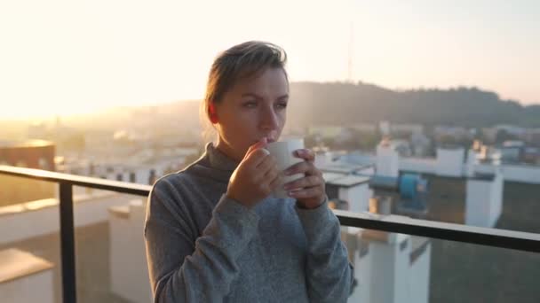 A nő egy csésze teával vagy kávéval kezdi a napját az erkélyen hajnalban. Városi táj a háttérben. Modern városi életmód - Felvétel, videó