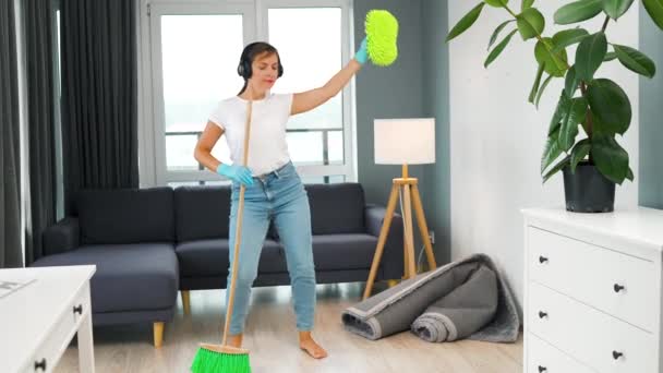 Blanke vrouw in koptelefoon schoonmaken van het huis en plezier hebben dansen met een bezem en washandje. - Video