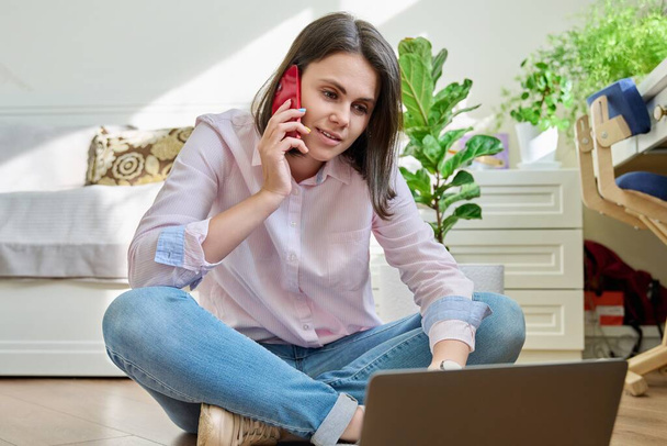 Young smiling woman sitting on floor at home talking on mobile phone using laptop. Education, study, remote online work, freelancing, youth concept - Valokuva, kuva