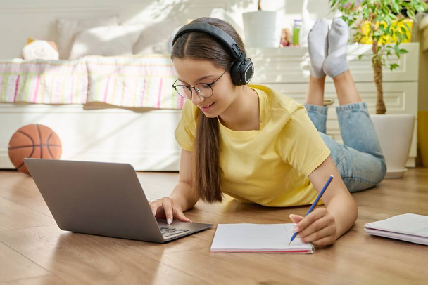 Adolescent teenage girl with glasses studying at home using laptop. Female with headphones and textbooks lying on floor in room. Education, adolescence, high school concept - Photo, image
