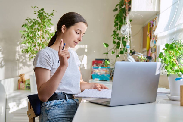 Teenage girl studying at home using laptop making notes in notebook. High school student preparing for control exam, doing homework, writing essay. Education, knowledge, adolescence concept - Foto, afbeelding