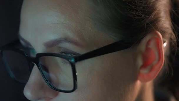 Woman in glasses looking on the monitor and surfing Internet at night. The monitor screen is reflected in the glasses. Work at night. Remote work. Extreme close-up - Materiał filmowy, wideo
