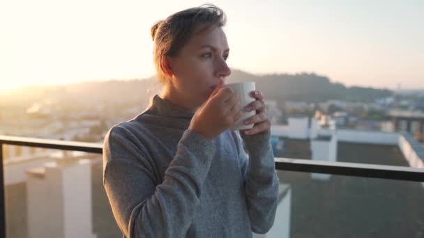 A nő egy csésze teával vagy kávéval kezdi a napját az erkélyen hajnalban, lassítva. Városi táj a háttérben. Modern városi életmód - Felvétel, videó