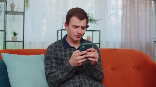 Teini-ikäisen aikuisen miehen muotokuva käyttää älypuhelimen kirjoittamista selaamalla, menettää yllättäviä äkillisiä lottotuloksia, huonoja uutisia, onnenmenetyksiä, pelin epäonnistumista. Nuori kaveri istuu oranssi sohvalla kotona olohuoneessa - Materiaali, video