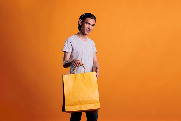 Happy smiling asian man showing shopping bag, store sale, bargain. Young asian shop client holding paper package, cheerful buyer carrying purchase, shopaholic buying products, - Photo, Image