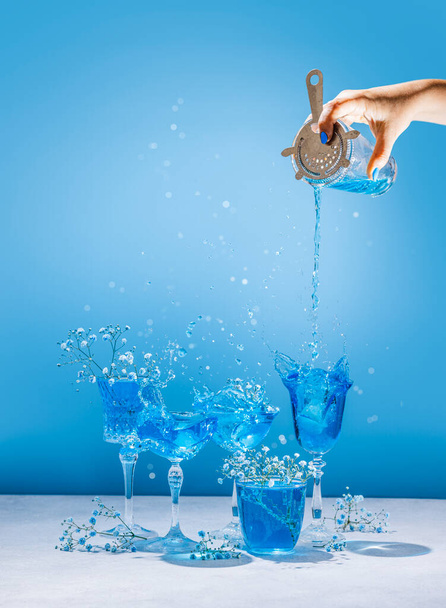 Diversi bicchieri di champagne eleganti con cocktail Blue Lagoon, mano delle donne sta versando cocktail da shaker a vetro coupé. Spruzzi, schizzi, fiori con gocce d'acqua sullo sfondo blu - Foto, immagini