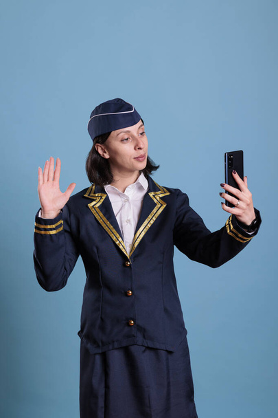 Flight attendant talking on smartphone videocall, holding mobile phone, having remote conversation, waving hi. Stewardess in uniform answering online teleconference call on telephone - Photo, Image