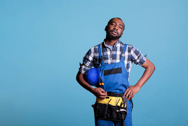 African american construction worker looking seriously at camera in studio shot against blue background. Construction worker at the end of the workday holding hard hat under arm. - Foto, afbeelding