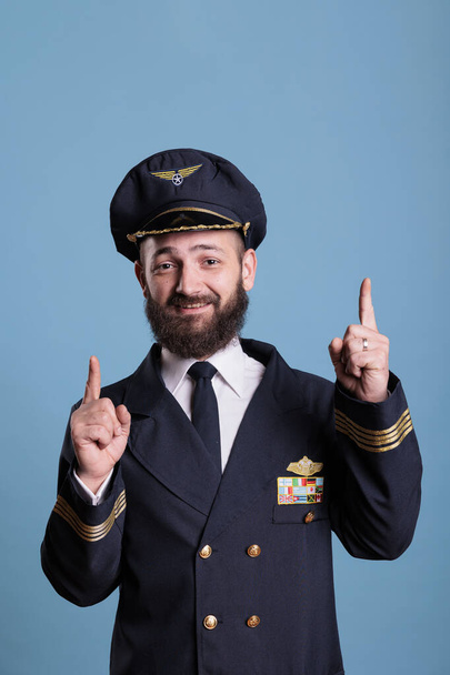 Πορτρέτο του χαμογελαστού αεροπόρος δείχνει επάνω με τα δάχτυλα δείκτη, διαφημιστικό προϊόν, φορώντας στολή της αεροπορίας. Αεροπλάνο πιλότος κοιτάζοντας κάμερα με forefingers δείχνει προς τα πάνω, χειρονομία προώθησης - Φωτογραφία, εικόνα