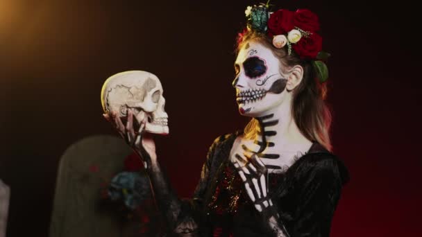 Horor Santa Muerte se dívá na lebku ve studiu, nosí strašidelný halloweenský kostým s čelenkou. Chovat se jako svatá okouzlující bytost na dios de los muertos, s černobílým make-upem. Ruční snímek. - Záběry, video