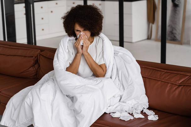 Beteg afrikai lány influenza fújja az orrát, és tüsszent a szalvéta, miközben ül kanapén otthon. Női láz, hideg és folyós orr takaróba csomagolva - Fotó, kép