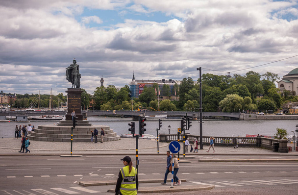 Švédsko, Stockholm - 16. července 2022: Cityscape, před královským palácem, kde se Skeppsbron setkává se Slottsbakenem u vody, stojí socha Gustava III pod modrým mrakem. Chodci přítomni - Fotografie, Obrázek