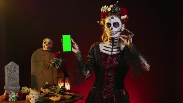 Strašidelný tradiční model držící zelenou obrazovku na smartphonu, pomocí mobilního telefonu s izolovanou mokup šablony. Žena oblečená jako Santa Muerte ukazující prázdné chroma klíč copyspace displej. - Záběry, video