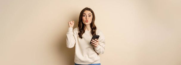 Happy girl holding smartphone and celebrating, winning on mobile phone app, standing over beige background. Copy space - 写真・画像