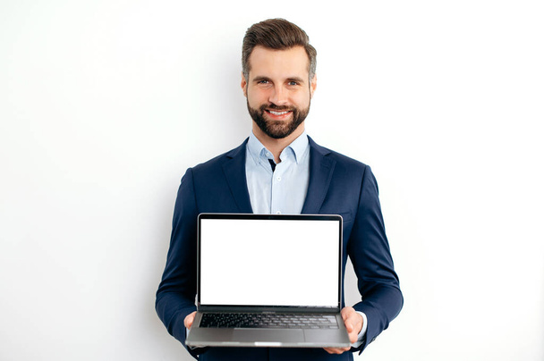 Advertisement concept. Positive caucasian bearded business man, in suit, programmer, IT specialist, seo, holds open laptop with empty mock-up screen in hand, stand on isolated white background, smiles - Foto, imagen