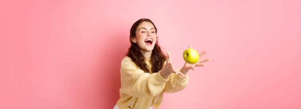 Feliz linda chica atrapa una manzana y se ríe, tiempo para un almuerzo saludable lleno de frutas, se levanta contra el fondo rosa. - Foto, imagen
