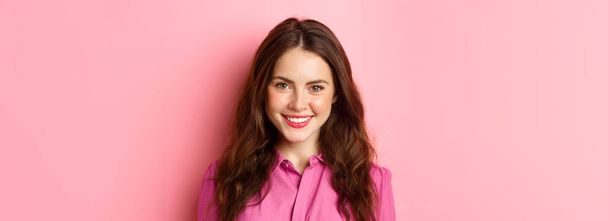 Close-up portret van stijlvolle moderne vrouw met make-up op en krullend kapsel, glimlachen en kijken vastberaden op de camera, staande tegen een roze achtergrond. - Foto, afbeelding
