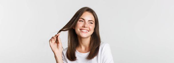 Portrét flirtování krásná dívka hraje s vlasy a usmívá se na kameru koketní, stojící bílé pozadí. - Fotografie, Obrázek