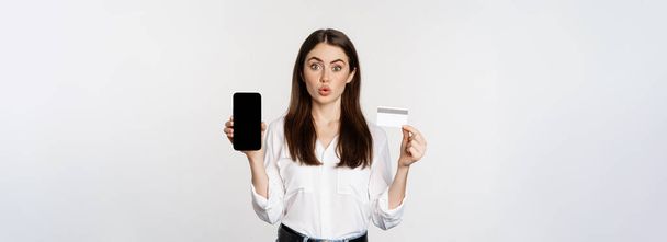Glückliche Frau zeigt Kreditkarte und Smartphone-Bildschirm, Konzept des Online-Shoppings, Kauf in App, steht vor weißem Hintergrund. - Foto, Bild