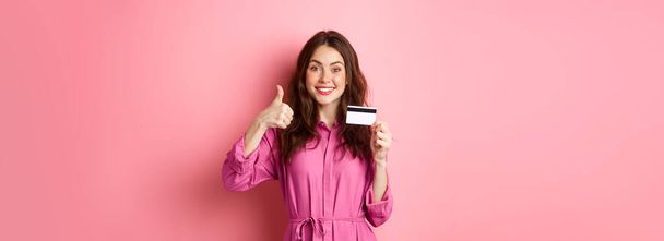 Chica morena mostrando los pulgares hacia arriba y sonriendo, recomendando banco, la celebración de la tarjeta de crédito de plástico, de pie sobre fondo rosa. Copiar espacio - Foto, Imagen