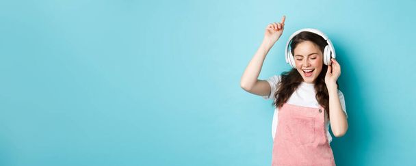 Κομψό κορίτσι της χιλιετίας που χορεύει με ανοιξιάτικο ντύσιμο, ακούει μουσική με ακουστικά και απολαμβάνει τον ήχο, στέκεται πάνω από το μπλε φόντο. - Φωτογραφία, εικόνα