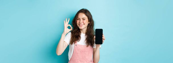 Velmi dobře. Usmívající se mladá žena zobrazující produkt on-line na prázdné obrazovce smartphone, doporučujeme vaši značku, aby ok znamení, stojí spokojeni s modrým pozadím. - Fotografie, Obrázek