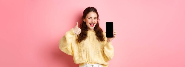 Дуже хороший додаток. Усміхнена задоволена жінка, яка показує великі пальці вгору і порожній екран смартфона, рекомендує додаток або веб-сайт, стоячи на рожевому фоні
. - Фото, зображення
