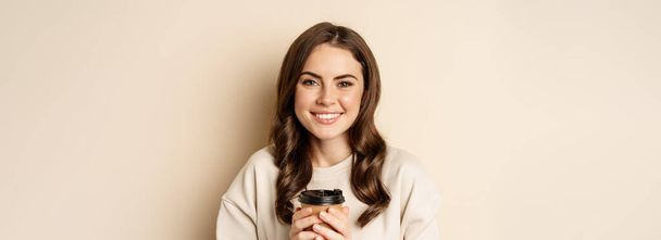 Beautiful authentic woman smiling, holding warm cup of coffee and looking happy, standing over beige background. - Foto, immagini