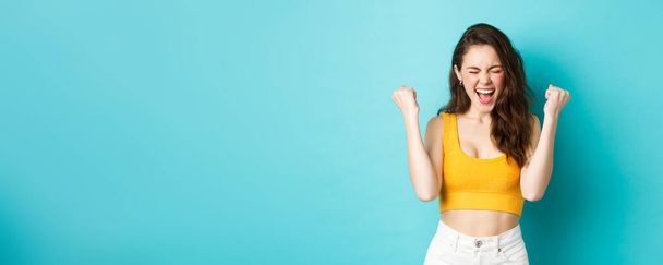 Summer holidays and emotions concept. Joyful girl shouting from satisfaction, showing fist pump and scream yes, achieve goal, celebrating victory or success, standing over blue background. - Photo, Image