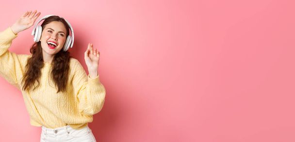 Ελκυστική στιλάτη γυναίκα που τραγουδάει το αγαπημένο της τραγούδι, ακούγοντας μουσική σε ασύρματα ακουστικά, χαμογελώντας και χορεύοντας, στέκεται πάνω από ροζ φόντο. - Φωτογραφία, εικόνα