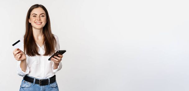 Smiling modern woman using credit card and mobile phone, paying, making purchase online, shopping on smartphone, standing over white background. - Fotó, kép